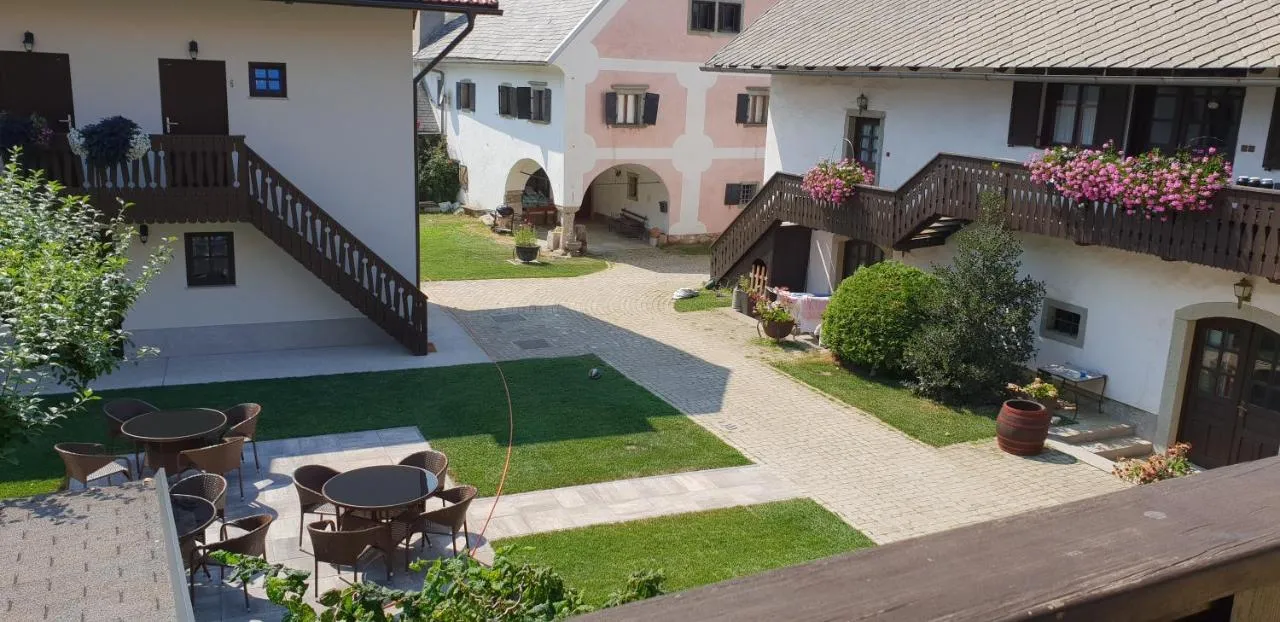 Dvorišče z galerije: mize na trati, leseno stopnišče, rožnata hiša z arkadami in rože na balkonih