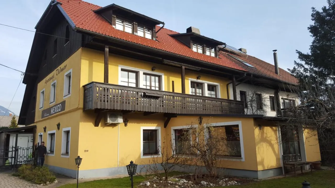 Rumena hiša z napisom Apartmaji Vidmar, lesenim balkonom in strešnimi frčadami